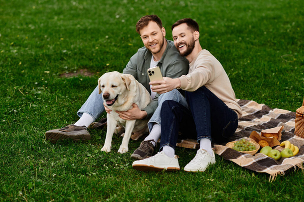 Δύο γενειοφόροι άνδρες απολαμβάνουν ένα πικνίκ με το λαμπραντόρ σκυλί τους σε ένα πράσινο πάρκο. Βγάζουν μια σέλφι με ένα τηλέφωνο και χαμογελούν.. - Φωτογραφία, εικόνα