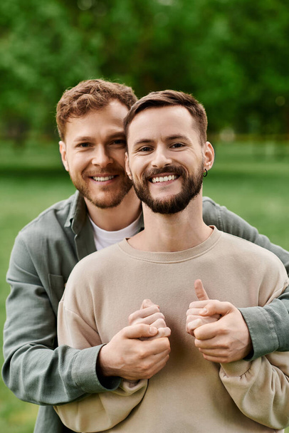 Two bearded men stand close, holding hands in a lush green park, showcasing their love and connection. - Photo, Image