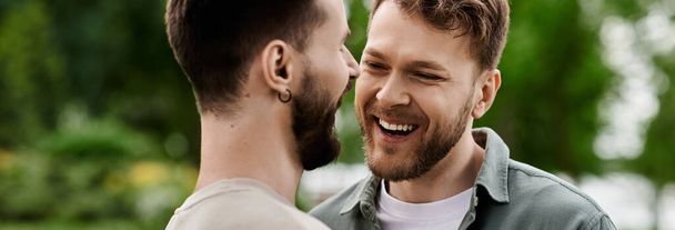 Dwóch brodatych mężczyzn śmieje się razem w bujnym zielonym parku, ciesząc się słonecznym dniem. - Zdjęcie, obraz