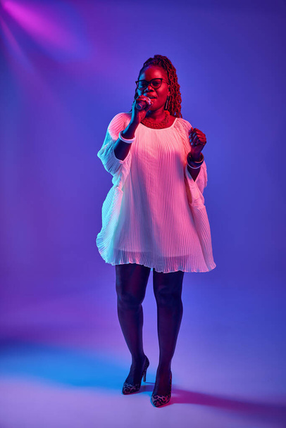 Mujer afroamericana en gafas actuando en el escenario, cantando en el micrófono en luz de neón sobre fondo degradado colorido. Concepto de música, festivales, arte contemporáneo, hobby, creatividad. Anuncio - Foto, Imagen