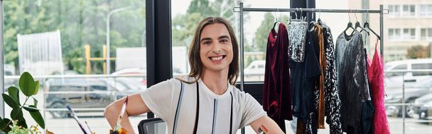 Ένας νεαρός γκέι άντρας χαμογελάει με αυτοπεποίθηση στο ατελιέ αποκατάστασης ρούχων του, περιτριγυρισμένος από ρούχα που περιμένουν μεταμόρφωση.. - Φωτογραφία, εικόνα