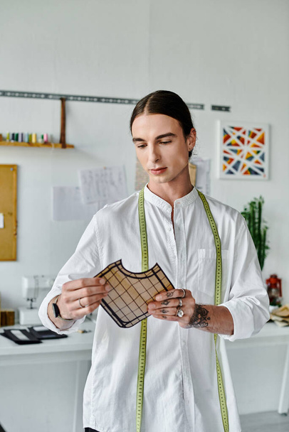 Um jovem de camisa branca, com uma fita métrica ao redor do pescoço, examina um pedaço de tecido em seu atelier de restauração de roupas.. - Foto, Imagem