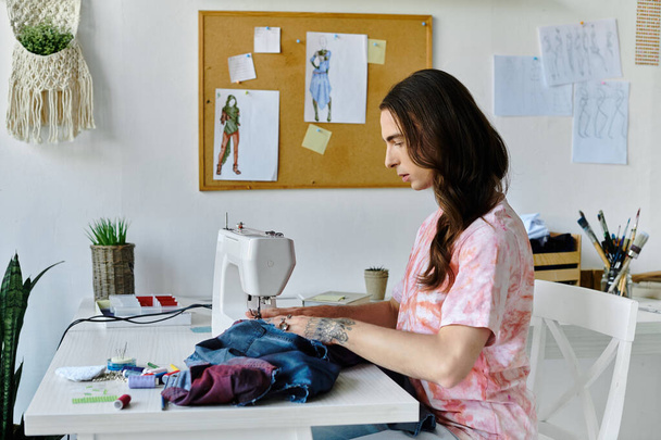 Молодой человек пришивает винтажную швейную машинку в своей домашней студии, окруженный красочными тканями и вдохновением. - Фото, изображение
