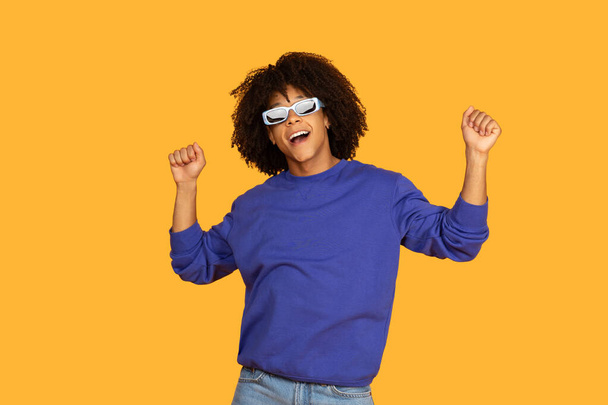 Um jovem alegre com cabelo encaracolado e danças de óculos de sol com as mãos levantadas contra um sólido fundo amarelo, vestindo uma camisola azul e jeans, expressando felicidade e energia despreocupada. - Foto, Imagem