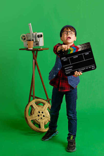 Opgetogen, klein kind, jongen verkleed als filmregisseur poserend met klapbord tegen groene studio achtergrond. Creatieve filmmaker. Concept van kinderdromen, toekomstige carrière, beroep, fantasie. Steun - Foto, afbeelding