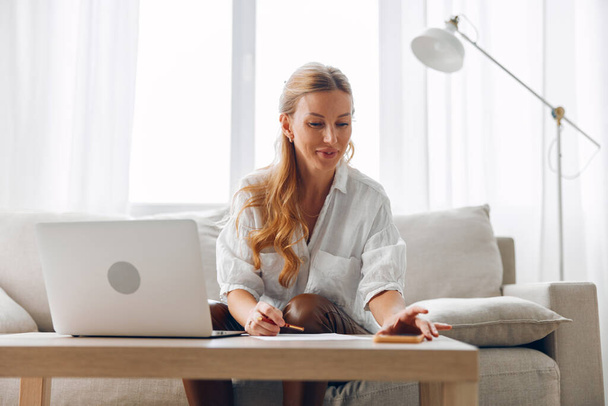 Vrouw die thuis werkt op de bank met laptop en pen, gericht op taak in gezellige woonkamer setting - Foto, afbeelding