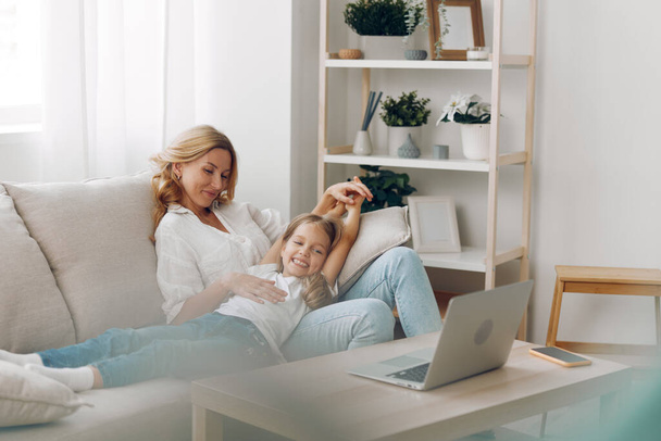 Mère et fille collant sur le canapé tout en utilisant un ordinateur portable, bras de femme enveloppé avec amour autour de l'enfant pour le soutien et le confort - Photo, image