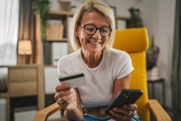 Ώριμη ηλικιωμένη γυναίκα αγοράζουν online με πιστωτική κάρτα και στο κινητό τηλέφωνο στο σπίτι - Φωτογραφία, εικόνα
