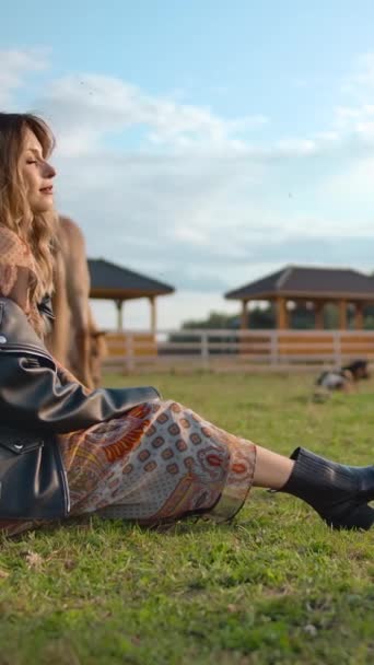 Fotoaparát se otáčí kolem šťastný fena ve stylovém oblečení sedí na trávě a upravuje bundu při trávení času v blízkosti koní na farmě. - Záběry, video