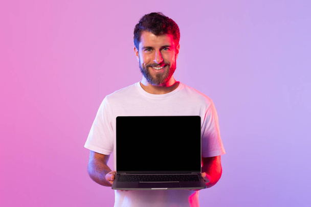 Młody mężczyzna w białej koszulce uśmiecha się trzymając w studio otwarty laptop z czarnym ekranem. Tło oferuje żywe różowe i fioletowe oświetlenie. - Zdjęcie, obraz