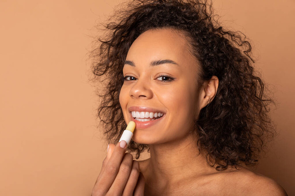 Deze foto toont een jonge Afro-Amerikaanse vrouw met krullend haar die lippenbalsem op haar lippen aanbrengt. Ze glimlacht, en haar uitdrukking is er een van geluk. De achtergrond is een warme bruine kleur. - Foto, afbeelding