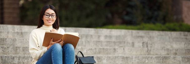 Una joven asiática con gafas se sienta en escalones de piedra frente a un edificio del campus. Ella está escribiendo en un cuaderno mientras mira hacia abajo en la página, copiar espacio - Foto, imagen