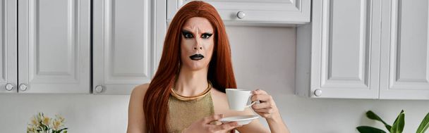 Королева трансвеститов в гламурной одежде наслаждается чашечкой кофе на кухне. - Фото, изображение