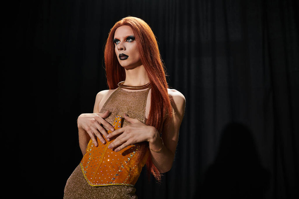 Czarująca drag queen w olśniewającym złotym i pomarańczowym stroju stoi na ciemnym tle. - Zdjęcie, obraz