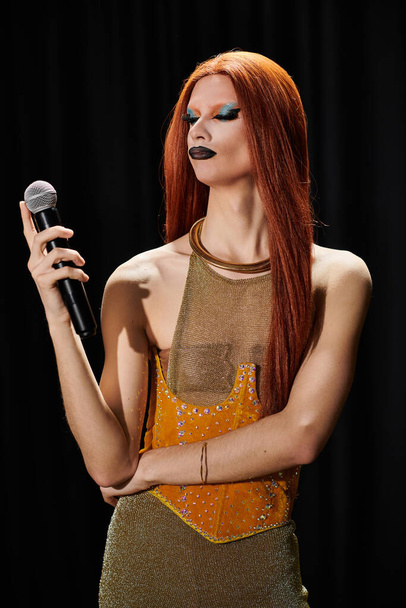 Una drag queen glamorosa con un deslumbrante atuendo dorado sostiene un micrófono. - Foto, imagen