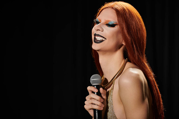 Una drag queen en un vestido de lentejuelas de oro sostiene un micrófono y se ríe. - Foto, Imagen