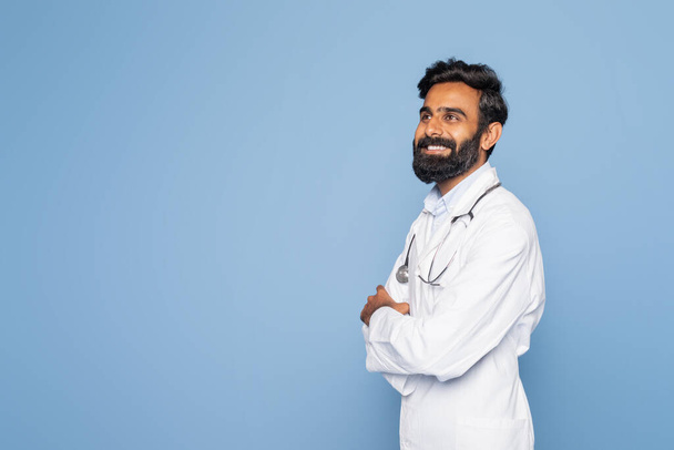 Бородатий індійський лікар у білому пальто та стетоскоп посміхається, впевнено стоїть на синьому фоні студії. Його руки схрещені, передають професіоналізм і тепло. - Фото, зображення
