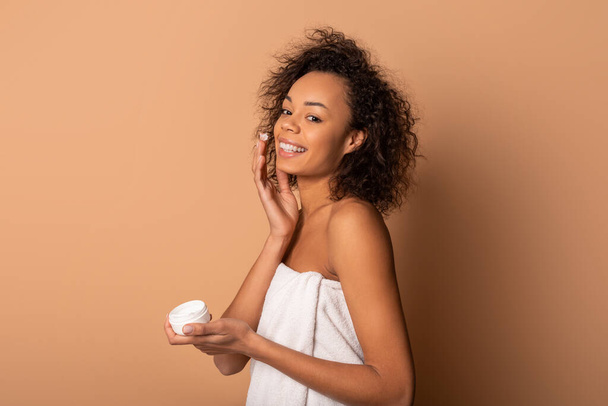 Afroamerikanische junge Frau mit lockigem Haar trägt nach dem Duschen Feuchtigkeitscreme auf ihre Haut auf. Sie trägt ein weißes Handtuch und lächelt in die Kamera. Hintergrund ist eine schlichte, hellbraune Wand. - Foto, Bild