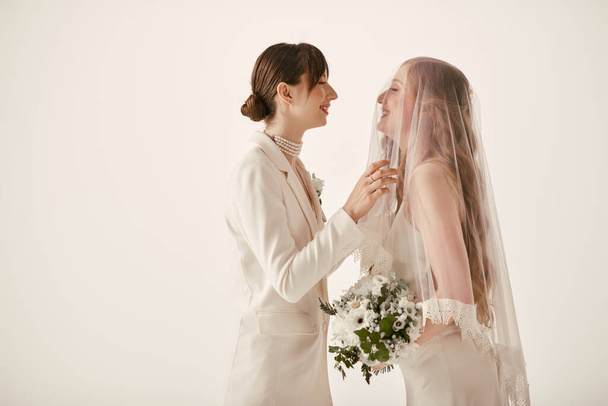 Zwei junge Frauen, beide in weißer Hochzeitskleidung, stehen einander gegenüber, lächeln und teilen einen liebevollen Moment während ihrer Hochzeitszeremonie. - Foto, Bild