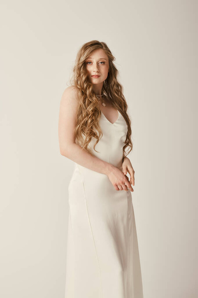 Eine schöne junge Braut im weißen Brautkleid steht vor weißem Hintergrund. Ihr langes, wallendes Haar trägt zu ihrer Eleganz bei. - Foto, Bild