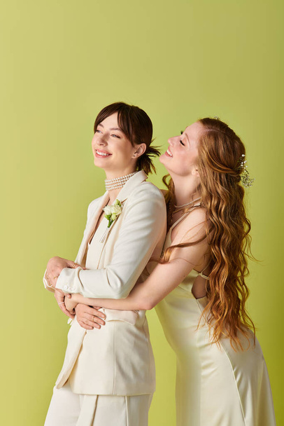 Two women in white wedding attire embrace each other against a green backdrop. - Fotoğraf, Görsel