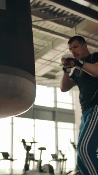 Boxer shadow boxing. Boxer swings and kicks and kicks the hanging punching bag. Bottom view. Modern fitness equipment and large windows in the background. Vertical shot. - Footage, Video