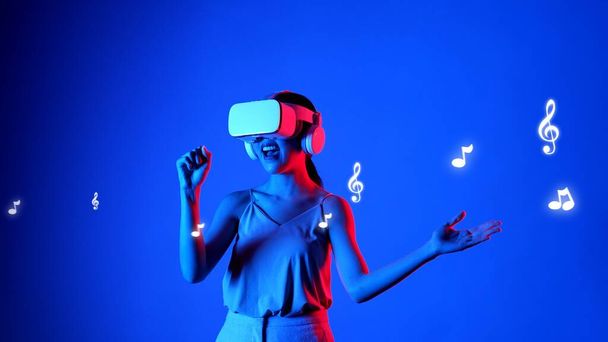 Mujer disfrutando cantando canción con auriculares VR con nota gráfica de neón melodía musical moderno diseño en estudio iluminación dinámica flotando en la música y la tecnología concepto fondo azul. Alucinaciones. - Foto, imagen