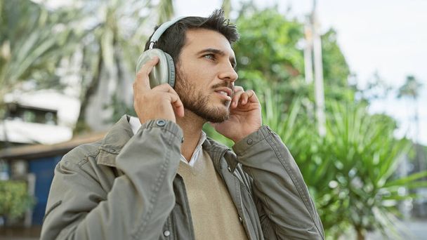 Hezký mladý hispánec s plnovousem má rád hudbu na sluchátkách venku v bujném městském parku. - Fotografie, Obrázek