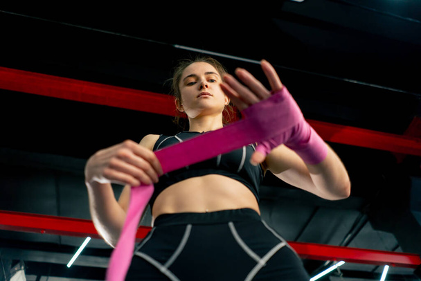 basso colpo a un club di boxe lotta una giovane ragazza con una felpa nera avvolge bendaggi elastici rosa per la boxe intorno alle mani preparandosi per l'allenamento - Foto, immagini