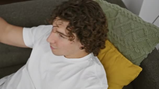 Nuori latino mies kiharat hiukset rentouttava sohvalla viihtyisässä olohuoneessa, hymyillen ja etsivät sisältöä. - Materiaali, video