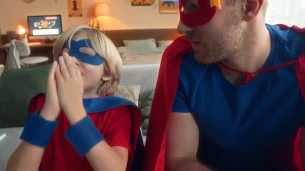 Excited Caucasian tween boy dressed in superhero costume throwing dice while playing boardgame with dad at home - Footage, Video