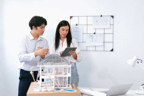 美しい若い慎重なプロジェクトマネージャーは,タブレットを使用して詳細を構築することをアジアのエンジニアに伝えますが,エンジニアはブループリントスキャッターで会議テーブル上の家モデルを測定します. インマキュライト - 写真・画像