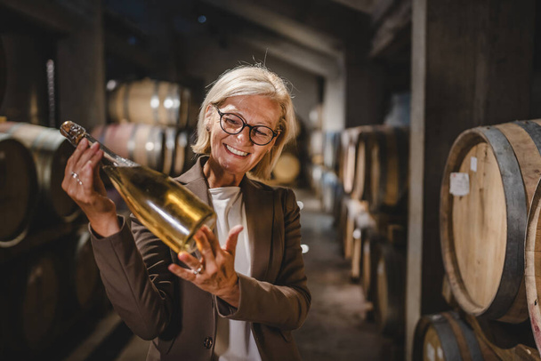 Érett idős nő borász állni az alagsorban tartsa üveg bor - Fotó, kép