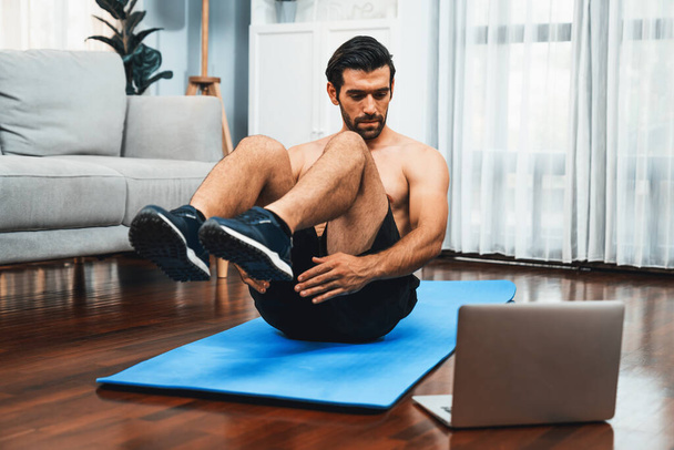 Athletic and sporty man doing crunch on fitness mat while follow online home workout exercise instruction for fit physique and healthy sport lifestyle at home. Online gaiety home exercise video. - Photo, Image