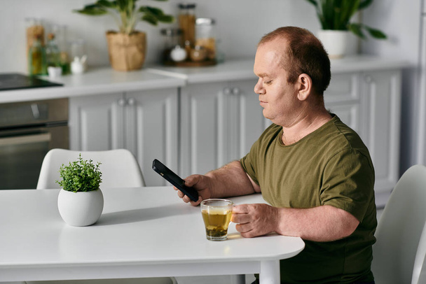 Mężczyzna z inkluzywnością w luźnych ubraniach siedzi przy stole w swoim domu, pijąc filiżankę herbaty, patrząc na telefon.. - Zdjęcie, obraz