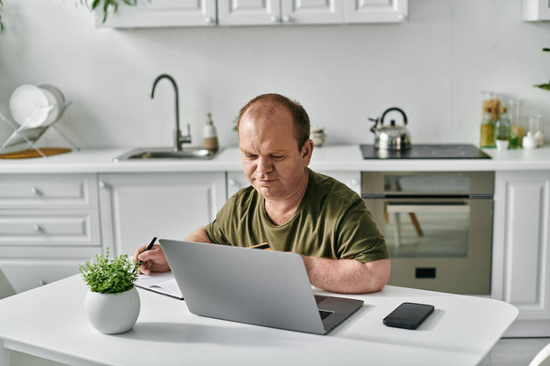 Un hombre con inclusividad en atuendo casual trabaja en su computadora portátil en una mesa en su cocina. - Foto, imagen