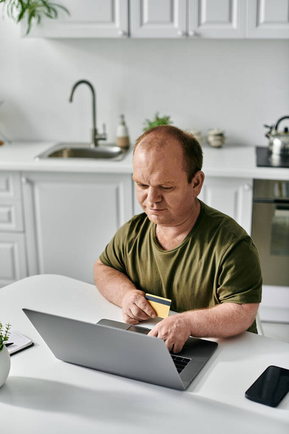 Um homem com inclusão senta-se em uma mesa de cozinha, usando seu laptop e um cartão de crédito para compras on-line. - Foto, Imagem