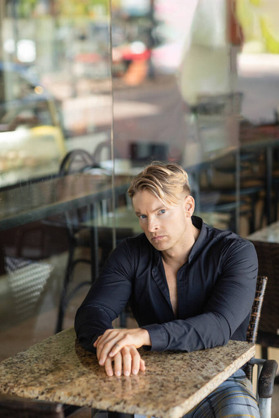 A handsome blonde man in a stylish black shirt sits at a table outside a cafe in Orlando, Florida, his gaze focused on something beyond the camera. - Фото, зображення