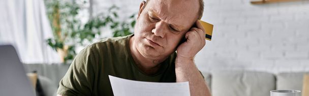 Egy zárkózott ember otthon ül, látszólag anyagi gondokkal terhelve, miközben egy dokumentumot vizsgál, és hitelkártyát tart magánál.. - Fotó, kép