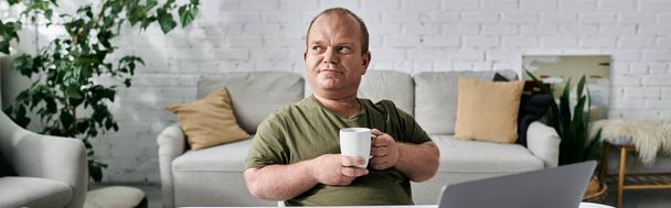 Un homme avec inclusivité s'assoit confortablement dans sa maison, tenant une tasse, regardant attentivement au loin. - Photo, image