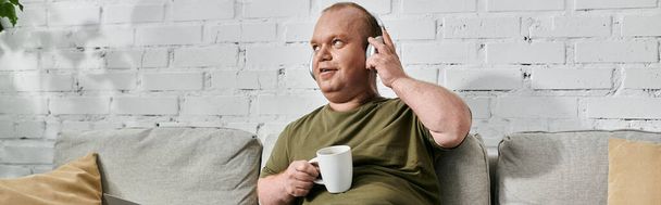 Egy inkluzivitású férfi fejhallgatót és alkalmi öltözéket visel, otthon pihen a kanapén, kezében egy csésze kávéval.. - Fotó, kép