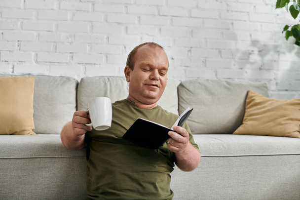 Un hombre con inclusividad se relaja en casa, disfrutando de una taza de café y un buen libro. - Foto, Imagen
