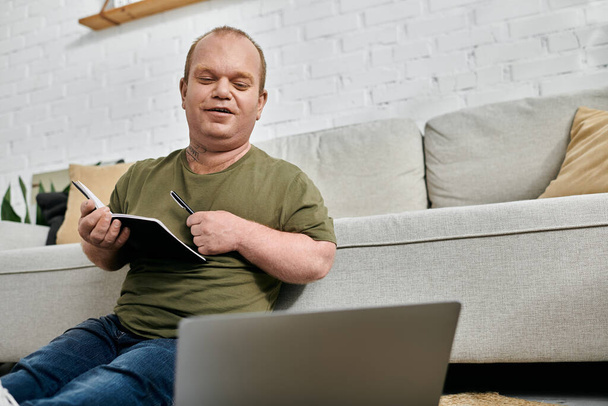 Чоловік з інклюзивністю сидить на дивані в своєму будинку, працює на ноутбуці і записує в блокноті. - Фото, зображення