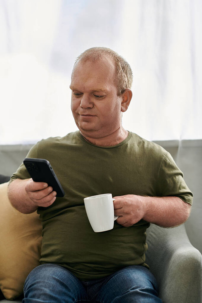 Ein Mann in Freizeitkleidung sitzt auf einem Sofa, in der einen Hand einen Kaffeebecher, in der anderen ein Smartphone.. - Foto, Bild