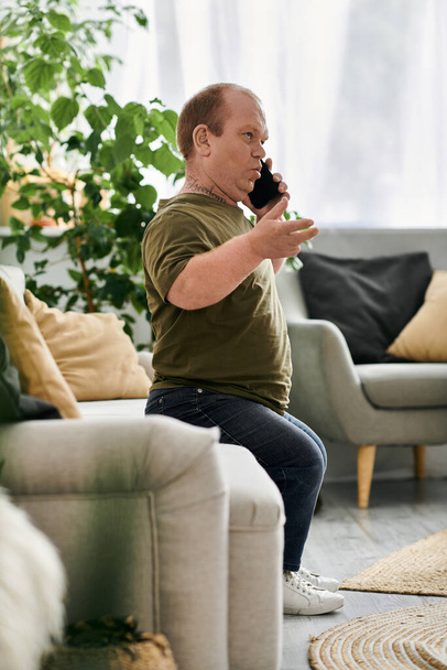 Egy inkluzivitású férfi ül egy kanapén a nappaliban, és telefonon beszél.. - Fotó, kép