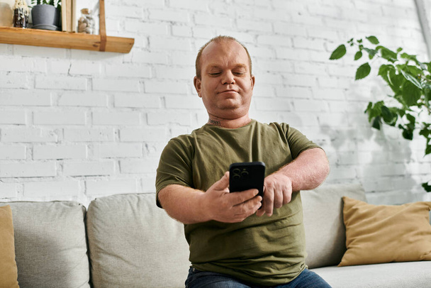 Mężczyzna z inkluzywnym ubiorem siedzi na kanapie w salonie i patrzy na telefon.. - Zdjęcie, obraz