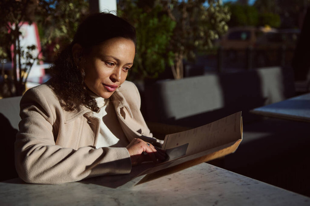 Een vrouw in een beige jas zit in een café een menu te lezen. Het zonlicht creëert een warme sfeer en benadrukt haar bedachtzame expressie. Bomen en tuinmeubilair zijn op de achtergrond te zien. - Foto, afbeelding