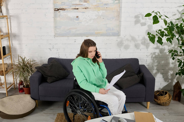 Egy barna nő ül egy tolószékben, telefonál és papírokat néz. Egy nappaliban van egy kanapéval és egy virággal a háttérben.. - Fotó, kép