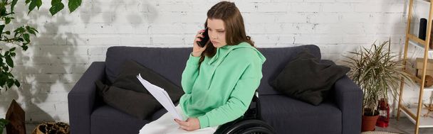 Молодая женщина с каштановыми волосами сидит в инвалидном кресле, разговаривая по телефону, держа в руках бумаги. She in her living room, surrounded by home furnishings. - Фото, изображение