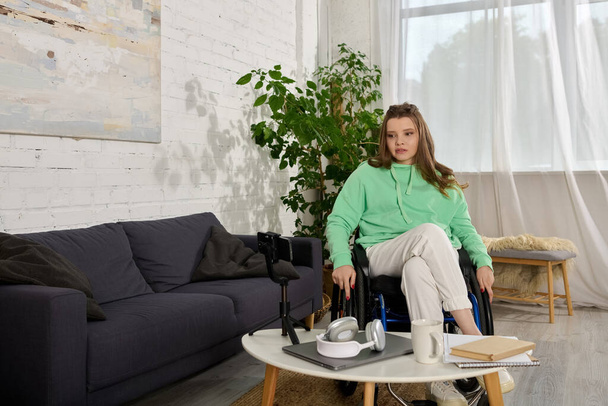 Una joven en silla de ruedas se sienta en su sala de estar, mirando el teléfono inteligente - Foto, Imagen